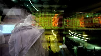 The image depicts a bustling financial trading environment, likely in a stock exchange. There are dynamic digital displays showing stock prices and market information, with green and red indicators reflecting changes in values. The foreground features figures in traditional attire, possibly indicating a cultural context. The overall scene conveys a sense of movement and activity, highlighting the fast-paced nature of trading. The use of motion blur adds to the dynamic feel of the environment.