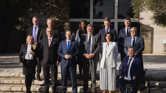 الصورة تظهر مجموعة من الأشخاص يقفون معًا في بيئة خارجية، ربما تكون حديقة أو منطقة أمامية لمبنى. يوجد في الخلفية مشهد طبيعي مع أشجار ونباتات. يرتدي الحضور ملابس رسمية وأنيقة، مما يشير إلى أن الصورة قد تم التقاطها في مناسبة رسمية أو مؤتمر. تتراوح تعبيرات الوجوه بين الابتسامات والجدية، مما يعكس جوًا من التعاون والتواصل.