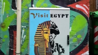 L'immagine mostra un armadietto metallico dipinto con un murale che presenta due volti distinti. A sinistra, c'è un'immagine stilizzata di un faraone, accompagnata da un invito a "Visit Egypt" e "Enjoy the". A destra, c'è un ritratto che sembra rappresentare un soldato o una figura autoritaria, con la parola "REGIME" sopra di esso. Lo sfondo è colorato e impreziosito da graffiti, creando un contrasto tra i due soggetti. La composizione sembra riflettere una critica sociale o politica legata all'Egitto.