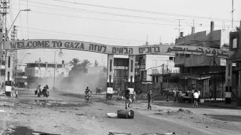 L'image montre une rue dans une ville, probablement à Gaza, avec une grande arche à l'entrée où il est écrit "Welcome to Gaza" en plusieurs langues. La scène est en noir et blanc, ce qui donne une atmosphère nostalgique ou dramatique. On aperçoit quelques personnes sur des bicyclettes et d'autres regroupées sur le côté de la route. Le décor urbain semble en désordre, avec des bâtiments qui montrent des signes d'usure et de dévastation. De la poussière s'élève, ajoutant à l'ambiance de la scène.