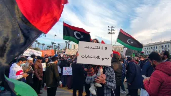 تظهر الصورة مشهداً من تظاهرة في مكان عام، حيث يحتشد الناس حاملين لافتات تتضمن عبارات تتعلق بالحرية والانتخابات. في الخلفية، ترفرف الأعلام الليبية بألوانها الأسود والأحمر والأخضر، مما يدل على الهوية الوطنية. يبدو أن الحشد يتنوع بين الرجال والنساء، وبعضهم يرتدي الكمامات، مما يشير إلى الحرص على الصحة العامة. السماء في الخلفية تظهر مع تزاحم الغيوم، مما يضيف جواً مميزاً إلى البيئة المحيطة بالتجمع.