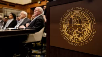 L'immagine mostra un incontro che si svolge in un ambiente formale, probabilmente in un'aula di tribunale. In primo piano si vede un simbolo dell'International Court of Justice (Corte Internazionale di Giustizia), con dettagli dorati che rappresentano l'autorità e la giustizia. Nel background si notano alcune persone sedute, molte delle quali sono visibili di spalle. L'atmosfera è seria e professionale, suggerendo un'importante deliberazione legale.