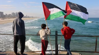 Nell'immagine, tre ragazzi sono in piedi su un molo che si affaccia su una spiaggia e sul mare. Due di loro tengono in alto delle bandiere palestinesi, che sono di colore nero, bianco, verde e rosso. Il ragazzo più piccolo, in una felpa grigia, sembra osservare attentamente l'orizzonte. La scena è illuminata da una luce naturale, con il cielo blu e alcune nuvole sparse. Le onde del mare si infrangono dolcemente sulla riva, creando un'atmosfera tranquilla. Gli altri due ragazzi indossano abbigliamento casual e sembrano condividere un momento di riflessione insieme.