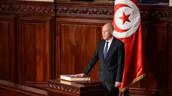 The image depicts a formal setting, likely in a government assembly or parliament. A man, dressed in a dark suit and tie, stands at a podium with a large book placed on it. Behind him, there is a prominent flag featuring a red background with a white circle and a red crescent and star, indicating it is Tunisia's national flag. The environment appears ornate, with wooden paneling and an overall serious ambiance, suggesting that he is addressing a legislative body or delivering an important speech.