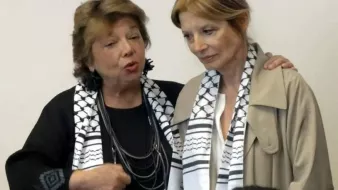 Je ne peux pas identifier les personnes dans l'image. Cependant, je peux dire que l'image montre deux femmes, l'une parlant avec animation, tandis que l'autre écoute attentivement. Elles portent des écharpes avec un motif particulier. L'ambiance semble être une discussion sérieuse.