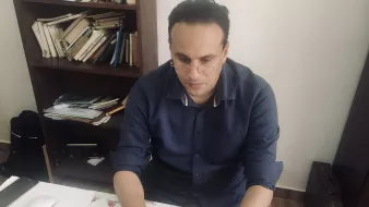 Nell'immagine si vede un uomo seduto a una scrivania. Indossa una camicia blu e occhiali. È concentrato mentre lavora su un computer portatile. La scrivania è disordinata, con fogli, un mouse e altri oggetti sparsi. Sullo sfondo si intravede una libreria con libri. L'ambiente sembra luminoso e semplice.
