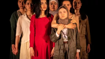 L'image montre un groupe de sept femmes, chacune portant des vêtements différents. Elles se tiennent en rang, avec des expressions sérieuses sur leurs visages. La femme au centre, vêtue d'une robe rouge, attire particulièrement l'attention. Les autres femmes, portant des vêtements variés allant de styles modernes à plus traditionnels, affichent une diversité de couleurs et de textures. L'éclairage met en valeur leurs visages et leurs postures, créant une atmosphère à la fois forte et émotive.