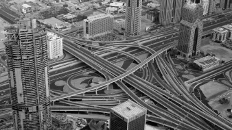 L'image présente une vue aérienne d'une métropole moderne, probablement Dubaï. On y voit de grandes tours élégantes et contemporaines, ainsi qu'un réseau complexe de routes et d'autoroutes qui s'entrelacent. Le contraste en noir et blanc accentue les formes géométriques des bâtiments et des routes, créant une impression de dynamisme et d'organisation. Des zones urbaines et dégagées suggèrent une planification urbaine sophistiquée.