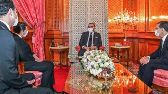 L'image montre un intérieur élégant avec un décor riche. Au centre, une table en verre repose sur un tapis rouge. On voit un homme assis, probablement un leader ou une personnalité importante, entouré de trois autres personnes, toutes en tenue formelle. Ils portent des masques, suggérant que la rencontre se déroule dans un contexte de précautions sanitaires. Le fond présente des murs ornés et un espace bien éclairé, avec des éléments décoratifs comme des fauteuils et des fleurs sur la table.