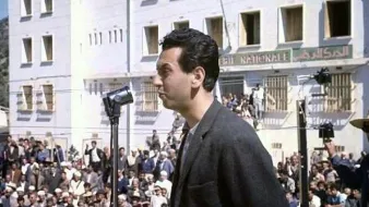 L'image montre un homme se tenant derrière un microphone, semblant s'adresser à une grande foule rassemblée devant lui. L'arrière-plan présente un bâtiment de style architectural simple avec plusieurs fenêtres, possiblement dans un contexte historique ou politique. Les membres de la foule semblent attentifs à son discours. L'éclairage naturel donne une ambiance claire à la scène, suggérant qu'elle pourrait avoir été prise en plein jour.
