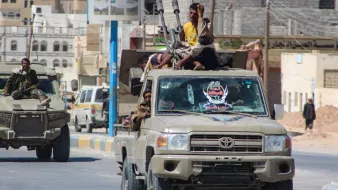 در این تصویر، یک خودروی نظامی تویوتا دیده می‌شود که بر روی آن چند نفر نشسته‌اند. خودروی نظامی دارای سلاح‌هایی است که روی آن نصب شده‌اند. در پس‌زمینه، ساختمان‌ها و خیابان‌های شهری قابل مشاهده هستند. وضعیت کلی به نظر می‌رسد که نشان دهنده یک وضعیت نظامی یا امنیتی باشد. نور روز و فضای خیابان نیز به تصویر اضافه شده است.