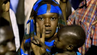 L'image montre une femme portant un foulard coloré, qui fait un signe de paix avec sa main. Elle tient un jeune enfant dans ses bras. L'arrière-plan semble flou, suggérant qu'elle se trouve dans une foule. Les expressions sur son visage et sa posture peuvent indiquer un moment de détermination ou de solidarité.