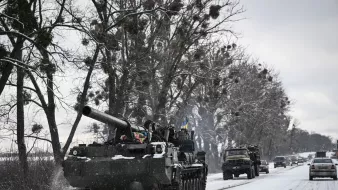 L'immagine mostra un carro armato che si muove lungo una strada innevata, circondato da alberi. Il carro armato ha una canna protrudente e presenta dettagli decorativi che potrebbero indicare l'appartenenza a una specifica unità militare. Accanto al carro armato, si intravedono anche alcuni veicoli, probabilmente militari, che viaggiano sulla stessa strada. Il paesaggio è caratterizzato da un'atmosfera invernale, con la neve che copre sia la strada che il terreno circostante.