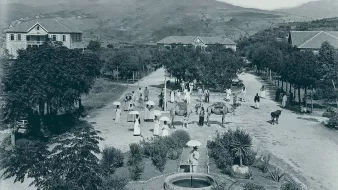 L'image semble représenter une scène rurale d'une époque passée. On peut voir un chemin central bordé d'arbres et de bâtiments à l'arrière-plan. Plusieurs personnes, vêtues de manière traditionnelle, se déplacent, certaines portant des parapluies. Des animaux comme des bœufs sont également présents, ajoutant à l'atmosphère agricole de la scène. Au premier plan, il y a un puits, probablement utilisé par les villageois. Les montagnes en arrière-plan complètent le paysage verdoyant. Cette image évoque une vie paisible et communautaire en harmonie avec la nature.