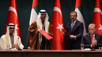 L'immagine mostra una cerimonia ufficiale con quattro uomini in abiti formali. Sullo sfondo si vedono le bandiere degli Emirati Arabi Uniti e della Turchia. Due degli uomini sono in piedi e sembrano scambiarsi documenti, mentre gli altri due sono seduti. L'atmosfera suggerisce un momento importante, probabilmente relativo a un accordo o a una collaborazione tra i due paesi.