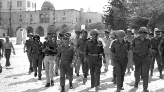 L'image montre un groupe de militaires marchant ensemble dans un environnement urbain. Ils portent des uniformes militaires et des casques. En arrière-plan, on peut voir des bâtiments avec une architecture typique, possiblement historique, ainsi qu'un dôme. L'atmosphère semble sérieuse, avec plusieurs soldats ayant une posture confiante. L'éclairage est lumineux, suggérant qu'il fait soleil. La scène donne une impression de mouvement et de coordination parmi les militaires.