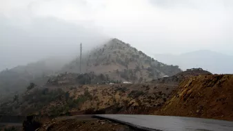 The image depicts a winding road leading through a mountainous landscape. In the background, a hill covered with low vegetation is partially obscured by mist or fog, creating a mysterious atmosphere. Atop the hill, there is a communication tower, and some vehicles are visible near the summit. The foreground features a dark, asphalt road that curves, suggesting a remote and rugged terrain. The overall scene conveys a sense of tranquility and solitude in nature.