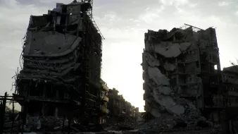 تظهر الصورة عمارتين محطمتين في منطقة يبدو أنها تعرضت لتدمير واسع. المباني مشوهة، حيث تظهر الجدران المنهارة والأسطح المدمرة، مما يعكس آثار الدمار. السماء في الخلفية تبدو ملبدة، مما يضيف شعورًا بالخيبة والخراب. المنطقة المحيطة يبدو أنها مهجورة، مما يعكس الأثر العميق للصراعات أو الكوارث التي أدت إلى هذا الوضع.