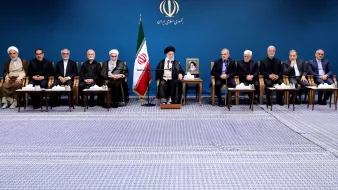 L'image montre un groupe de personnes assises en rangée sur un tapis, dans un environnement formel. En arrière-plan, il y a un drapeau iranien et un portrait d'une figure importante, probablement un leader. Les participants semblent être des hommes en costume sombre et en tenue religieuse, certains portant des turbans. Ils semblent engagés dans une discussion ou une réunion officielle. L'ambiance est solennelle et professionnelle.