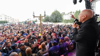 L'image montre un homme, probablement un orateur, s'adressant à une grande foule de personnes. La scène semble se dérouler à l'extérieur, avec des arbres en arrière-plan. Le public est très nombreux et semble attentif à ce qui est dit. À l'avant de la scène, des membres des forces de sécurité ou des agents en uniforme sont présents pour assurer l'ordre. L'atmosphère paraît dynamique et engagée, suggérant un événement public important.