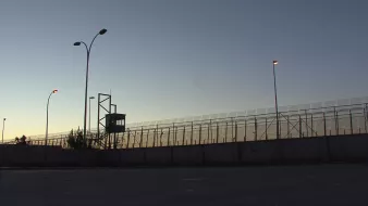 L'image montre une clôture de sécurité, probablement d'une prison ou d'une zone contrôlée. On aperçoit une tour d'observation au fond, illuminée par des lampadaires. La scène se déroule au crépuscule, avec un ciel qui passe des teintes orange et bleues, créant une atmosphère à la fois calme et tendue. Le mur en béton à la base de la clôture souligne le caractère restrictif de l'endroit.