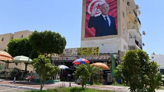 در این تصویر، یک دیوار بزرگ ساختمان نشان داده شده است که بر روی آن تصویری از یک فرد با اشاره به سمت بیننده مشاهده می‌شود. این شخص لباس رسمی به تن دارد و در پس‌زمینه تصویر، پرچم تونس دیده می‌شود. در پایین تصویر، یک کارگاه یا فروشگاه کوچک قرار دارد که چند نفر در زیر چادرهای رنگی نشسته‌اند. درختان سبز و دوچرخه‌ها نیز در تصویر وجود دارند. آسمان آبی و صاف به نظر می‌رسد.