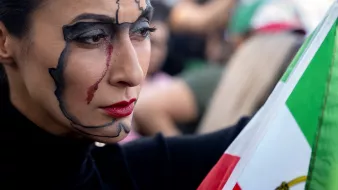 L'immagine mostra una donna con un trucco facciale elaborato, che rappresenta una mappa del suo paese. Ha un'espressione seria e intensa, con un tratto rosso che scende lungo il suo viso, suggerendo emozione o una narrazione profonda. In una mano tiene una bandiera, evidenziando un forte senso di identità nazionale e di protesta. Sullo sfondo si intravedono altre persone, creando un'atmosfera di raduno o manifestazione.
