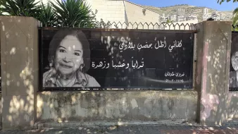 L'image montre une grande affiche accrochée à une clôture. Elle présente un portrait en noir et blanc d'une femme souriante. À côté de son visage, on trouve une citation en arabe, ainsi qu'un nom et une date en bas. L'arrière-plan montre des bâtiments et des végétaux, indiquant que l'affiche est placée dans un environnement urbain. L'ensemble véhicule une ambiance d'hommage ou de souvenir.