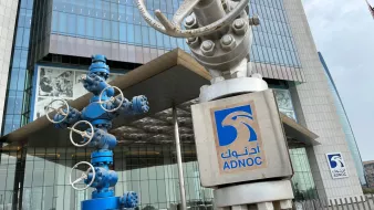 تصویر نشان‌دهنده نمای ساختمان شرکت نفت ابوظبی (ADNOC) است. در جلو، دو ساختار فلزی وجود دارد که شبیه به تجهیزات صنعتی و دکل‌های نفتی هستند. یکی از این تجهیزات به رنگ آبی و دیگری نقره‌ای است. در سمت راست تصویر، لوگوی ADNOC با نوشته عربی و انگلیسی قابل مشاهده است. ساختمان دارای ظاهری مدرن و شیشه‌ای است که عناصر طراحی معاصر را به نمایش می‌گذارد.