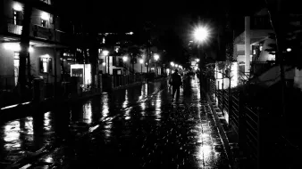 L'image montre une rue mouillée de nuit, éclairée par des lampadaires. Le sol reflète la lumière, créant un effet brillant. On peut voir quelques personnes marchant sur le trottoir, tandis que des bâtiments se dressent de chaque côté, illuminés par des lumières. L'atmosphère semble calme et sereine, typique d'une soirée de pluie.