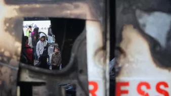 تصویر نشان‌دهنده‌ی یک صحنه‌ی غم‌انگیز است که در آن چندین کودک و بزرگسال درحال مشاهده‌ی یک وضعیت بحرانی به نظر می‌رسند. در پس‌زمینه، یک وسیله نقلیه آسیب‌دیده دیده می‌شود که حالت سوخته و خراب دارد. نام "پرس" بر روی آن به وضوح مشاهده می‌شود. در داخل نمای این وسیله، افرادی با حالت‌هایی جدی و نگران دیده می‌شوند که شاید به دنبال کمک یا راهی برای فرار از وضعیت خطرناک باشند. احساس تنش و اضطراب در این تصویر محسوس است.