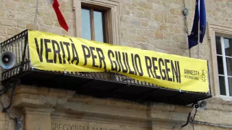 L'immagine mostra un grande striscione giallo appeso a un balcone di un edificio. Sullos striscione è scritto "VERITÀ PER GIULIO REGENI", accompagnato dal logo di Amnesty International. Nel contesto, si possono notare delle bandiere, probabilmente italiane, che sventolano. L'atmosfera suggerisce una manifestazione di sostegno e richiesta di giustizia per Giulio Regeni, un caso di rilevanza pubblica.