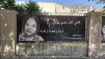 L'immagine mostra un grande striscione che riporta una fotografia di una donna sorridente, accompagnata da un testo in arabo. Il contesto sembra essere un'area urbana, con edifici sullo sfondo e piante verdi nelle vicinanze. L'atmosfera è accogliente e il messaggio sullo striscione appare positivo e invitante.
