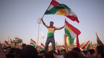L'image montre une grande foule célébrant, probablement lors d'un événement culturel ou politique. Au premier plan, un homme se tient debout sur une autre personne, tenant des drapeaux kurdes. Il porte un maillot aux couleurs du drapeau kurde, et son expression semble joyeuse et enthousiaste. En arrière-plan, on peut voir de nombreuses autres personnes agitant également des drapeaux, créant une atmosphère festive et énergétique. Le ciel est clair, ce qui ajoute à l'ambiance dégagée de la scène.