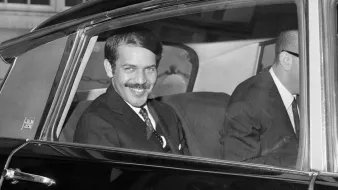 The image shows a man sitting in the back seat of a car, smiling at the camera. He is dressed in a formal suit and tie, with his hair neatly styled. Another person, partially visible, is seated next to him, also dressed in a suit. The setting appears to be a city, indicated by buildings in the background. The photograph has a classic black-and-white aesthetic, suggesting it was taken several decades ago.