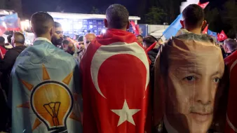 L'immagine mostra un gruppo di persone che indossano mantelli colorati. Due di loro hanno i mantelli decorati con il simbolo del partito AKP (Partito della Giustizia e dello Sviluppo) e la bandiera turca. Inoltre, uno dei mantelli presenta l'immagine di una figura pubblica, probabilmente un leader politico turco. Sullo sfondo si possono vedere altre bandiere e un'atmosfera di festa o celebrazione.