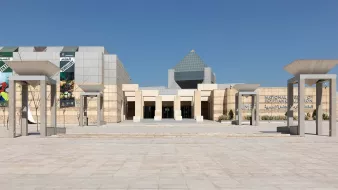 L'image montre l'entrée d'un bâtiment moderne, probablement un musée ou un centre culturel, avec une architecture distinctive. En façade, on voit plusieurs colonnes soutenant un toit en forme de voile, et une grande structure pyramidale se dresse à l'arrière. Les murs sont en pierre beige, donnant un aspect élégant et contemporain. Le ciel est clair, sans nuages, ce qui crée une atmosphère lumineuse et accueillante. Les éléments graphiques sur les murs suggèrent des expositions ou des événements.