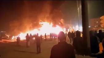 L'image montre une scène nocturne où un bâtiment est enflammé, créant une grande colonne de feu et de fumée. Des personnes se tiennent au premier plan, observant la situation. On peut noter une ambiance de tension, avec des lumières des bâtiments environnants qui ajoutent à l'atmosphère dramatique. Les silhouettes des personnes sont découpées contre la lumière des flammes.