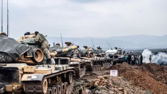 L'image montre une ligne de chars militaires, probablement en position de déploiement ou d'entraînement. Les chars sont recouverts de bâches et semblent être stationnés sur un terrain accidenté, entourés de collines. Au loin, on peut apercevoir des militaires rassemblés autour de ce qui semble être un poste de commandement ou d'observation. Il y a également des signes ou des marquages au sol, et l'atmosphère paraît chargée, avec une légère brume ou fumée dans l'air.