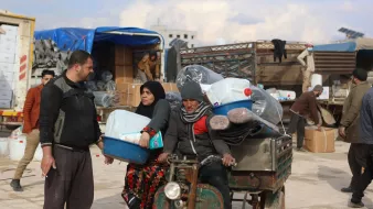 Nell'immagine si vede una scena di un'area di stoccaggio o mercato. Ci sono alcune persone che sembrano occupate a scaricare e trasportare materiali. Un uomo e una donna sono in primo piano: l'uomo indossa abiti scuri e tiene una cassetta, mentre la donna è vestita con un lungo mantello scuro e apparente una certa preoccupazione sul volto. Dietro di loro, ci sono merci come bottiglie di plastica e tappeti, oltre a veicoli e altri uomini che svolgono attività di carico e scarico. L'atmosfera sembra essere quella di un'operazione di supporto umanitario.