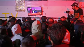 در تصویر، گروهی از افراد در حال تماشای تلویزیونی هستند که بر روی دیواری نصب شده است. تلویزیون یک برنامه خبری را نمایش می‌دهد و یک سخنران در حال صحبت است. افراد به سمت صفحه تلویزیون خیره شده‌اند و جو عمومی تصویر نشان‌دهنده توجه و کنجکاوی آن‌هاست. فضای اطراف کمی تاریک و نور محیطی کم است، به طوری که نور تلویزیون بیشتر مشخص است. برخی از افراد در لباس‌های گرم، نظیر هودی و کلاه، حضور دارند. یکی از افراد در سمت راست با دوربین در حال عکاسی است.