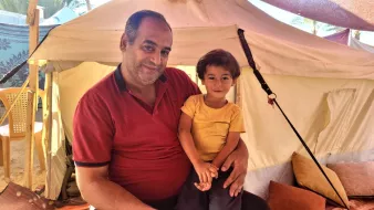Nell'immagine si vede un uomo sorridente che tiene in braccio un bambino. L'uomo indossa una maglietta rossa e il bambino ha una maglietta gialla. Sono entrambi all'interno di una tenda, circondati da cuscini e altri oggetti. L'atmosfera sembra calda e accogliente, con una luce naturale che illumina la scena.