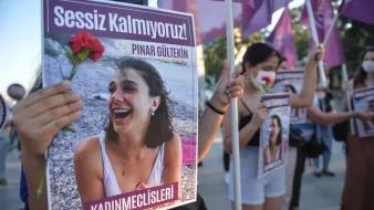 تظهر الصورة مجموعة من النساء أثناء تظاهرهن. تحمل إحداهن لافتة مكتوب عليها "Sessiz Kalmıyoruz!" (لا نصمت!) وصورة لامرأة تُدعى "Pınar Gültekin". كما تحمل إحدى المتظاهرات زهرة حمراء، مما يشير إلى أن المظاهرة تتعلق بقضية اجتماعية أو حقوق النساء. يُظهر المشهد روح التضامن والمطالبة بالعدالة.