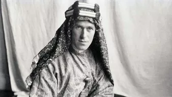 L'image montre un homme portant un costume traditionnel. Il est vêtu d'une tunique ornée et d'un headdress décoratif. Son regard est sérieux et il tient un livre dans ses mains. L'arrière-plan est sobre, ce qui met en valeur son apparence et son habillement. La photo semble être ancienne, ce qui lui donne un aspect historique.