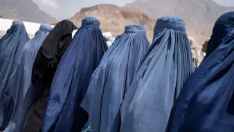 در این تصویر تعدادی زنان دیده می‌شوند که پوشش‌هایی به رنگ‌های آبی و مشکی بر تن دارند. آنها از پشت به دوربین ایستاده‌اند و فقط قسمت‌هایی از پوشش‌هایشان قابل مشاهده است. نمای背景 کوه‌ها و فضای باز است، که ممکن است به منطقه‌ای کوهستانی اشاره داشته باشد. این تصویر حس سنتی و فرهنگی خاصی را منتقل می‌کند.
