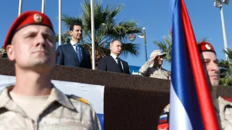 The image depicts a military ceremony or event, featuring several individuals of importance. In the foreground, soldiers in military uniforms with red berets are standing at attention. In the background, a group of officials is present, including two prominent figures positioned together: one in a suit and tie and the other in military attire. There's a large flag in the foreground, possibly representing a nation involved in the event. The setting appears outdoors, with palm trees visible, suggesting a warm climate. The overall atmosphere seems formal and significant, likely indicative of a diplomatic or military alliance.
