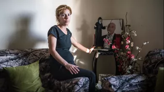 L'image montre une femme assise sur un canapé, portant une tenue noire. Elle est positionnée dans une pièce au mur neutre, avec un éclairage doux. À côté d'elle, il y a un petit meuble sur lequel se trouvent des objets décoratifs, ainsi qu'une photo encadrée d'un homme. Le décor du canapé est à motifs camouflage, et quelques fleurs artificielles ajoutent une touche de couleur à l'environnement. L'expression de la femme semble sérieuse, reflétant peut-être une émotion ou une réflexion profonde.