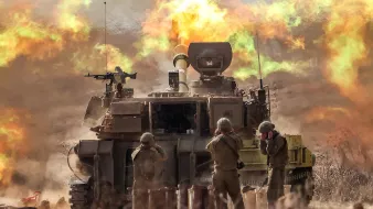 L'immagine mostra un carro armato in azione, con un cannone montato che spara. Sullo sfondo si vede un'esplosione che genera fumi colorati, creando un'atmosfera drammatica. Davanti al carro armato, alcuni soldati sono in posizione, probabilmente impegnati nell'operazione militare. La scena è caratterizzata da polvere sollevata e un'illuminazione intensa, evidenziando l'intensità dell'azione.