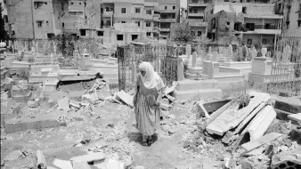L'image montre une femme vêtue d'un long habit, marchant au milieu d'un cimetière. Le sol est désordonné, avec des débris éparpillés, ce qui suggère qu'il y a eu des destructions récentes dans la zone. En arrière-plan, on peut voir des bâtiments, probablement endommagés, qui entourent le cimetière. L'atmosphère paraît lourde, évoquant des sentiments de tristesse et de désolation.