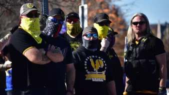 The image depicts a group of people standing together, wearing clothing that features dark colors and some yellow accents. Many of them have their faces covered with bandanas or masks, and they are wearing sunglasses. Their expressions and postures suggest a sense of solidarity or confrontation. The group is likely gathered for a specific event or rally, as indicated by their coordinated appearance. In the background, there are trees and other people visible, suggesting an outdoor setting.